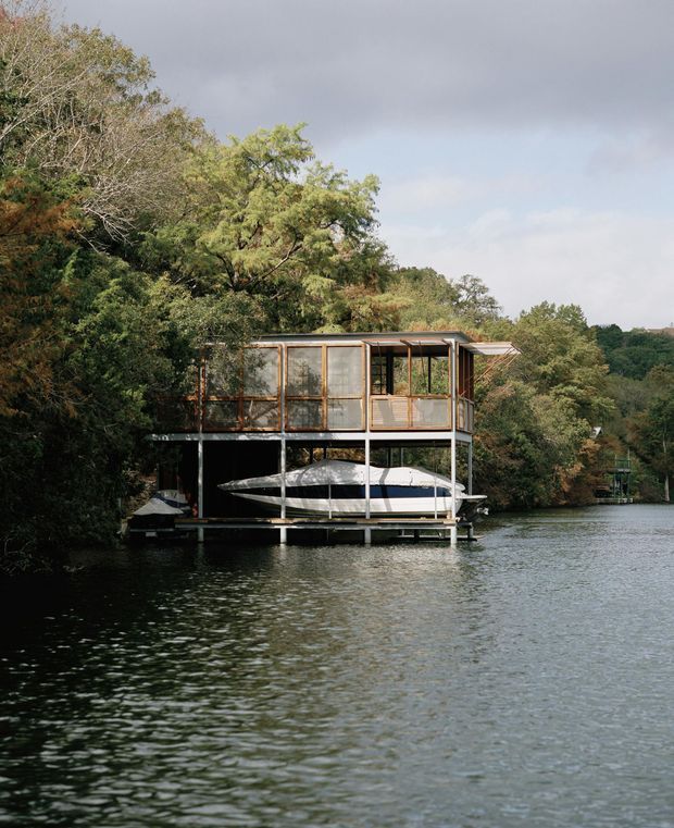 A compact elevated waterside structure sits on a floating or stilted platform at the edge of a tree-lined lake. The upper level is enclosed with extensive glazing and framed in warm timber, while the open lower level shelters a long boat. The scene is photographed from the water under an overcast sky, with dense natural vegetation surrounding the structure.