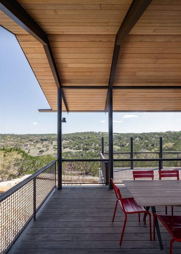 A covered outdoor dining deck overlooking a rugged, tree-covered landscape. The terrace features dark timber decking, a timber-lined ceiling with exposed black steel beams and posts, black wire-mesh balustrades, and a dark dining table with bright red chairs. The open-sided structure creates a sheltered yet panoramic outdoor setting.
