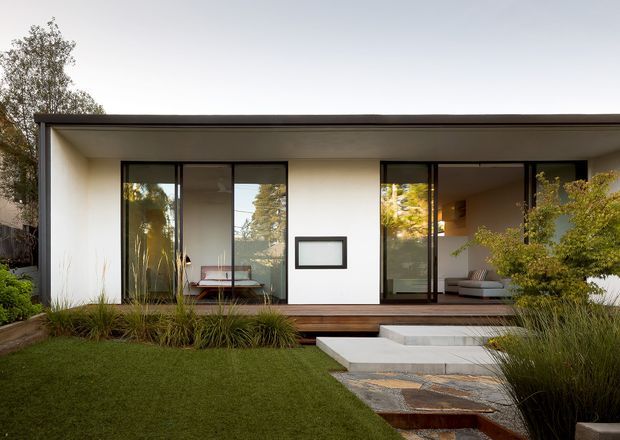 A single-storey contemporary residence with a flat roof, crisp white walls, dark window and door frames, and extensive floor-to-ceiling glazing. The house opens onto a landscaped front garden with clipped lawn, ornamental grasses, shrubs, broad concrete steps, and a layered stone pathway. Interior furnishings are visible through the glazing, including a dining area and lounge seating.