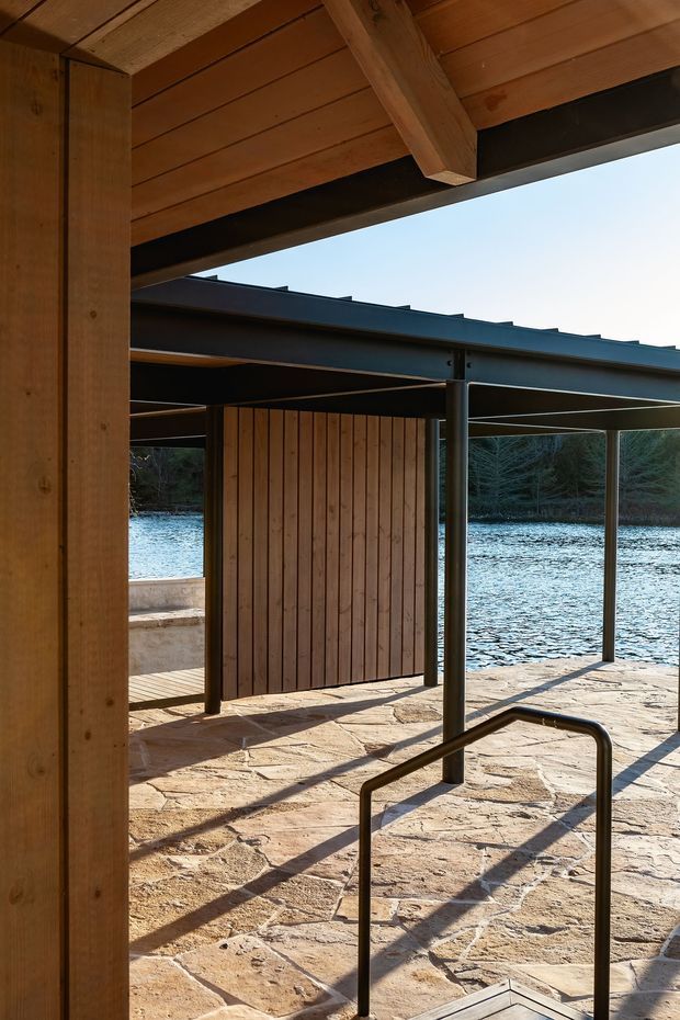 A covered lakeside outdoor structure with warm timber posts and ceiling, dark steel framing, vertical timber screening, and a stone-paved terrace descending toward the water. A simple dark metal handrail and covered platforms create a contemporary waterfront access area, with trees and the lake visible beyond.