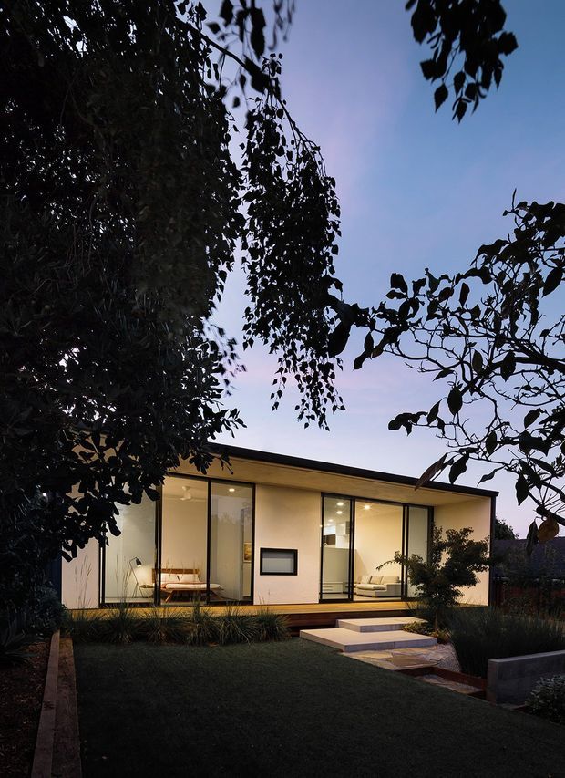 Dusk exterior view of a compact modern single-storey house set within a landscaped garden. The home has a simple low-pitched roof, pale rendered walls, extensive floor-to-ceiling glazing with dark slender frames, and a timber deck reached by broad pale steps. Warm interior lighting reveals living spaces through the windows, while mature planting frames the foreground.