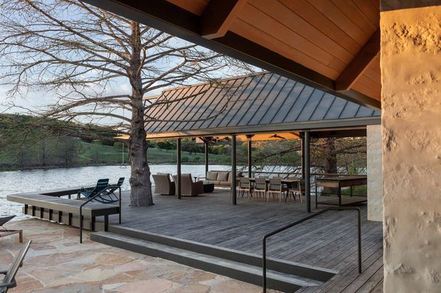 A covered outdoor entertaining deck beside a lake, framed by a warm timber-lined roof and natural plaster wall. The expansive timber decking includes lounge and dining furniture, a built-in outdoor kitchen or bar area, and steps leading to a stone-paved terrace. A separate pavilion with a dark standing-seam metal roof shelters the seating area, while mature trees and the water form the background.