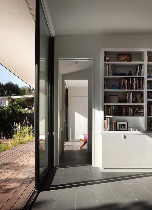 A bright, contemporary interior corridor or living area with full-height dark-framed glazing opening onto a timber deck and landscaped garden. The room has a grey tiled floor, pale grey walls and ceiling, and extensive built-in white shelving and lower cabinetry filled with books and display objects. A bedroom is visible through the doorway and reflection, with sunlight creating strong linear shadows across the floor.