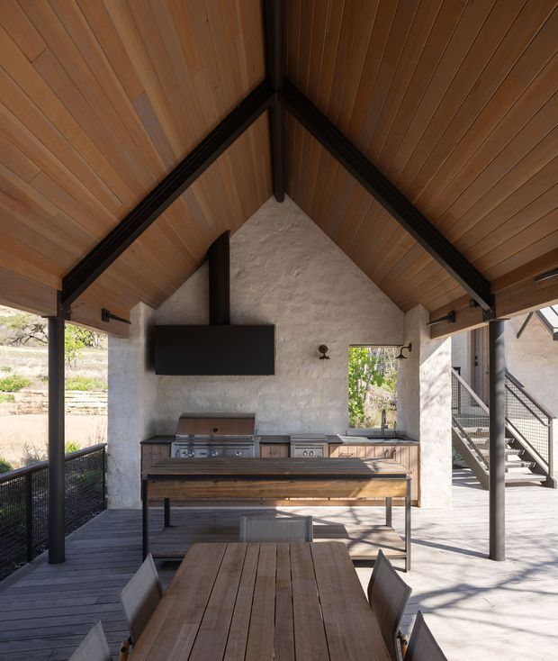 Covered outdoor entertaining area with a pitched timber-lined ceiling, exposed dark structural beams, textured light-rendered walls, built-in barbecue cabinetry and a large timber dining table. Open sides connect the space to a deck and exterior stairs, creating a refined indoor-outdoor kitchen and dining setting.