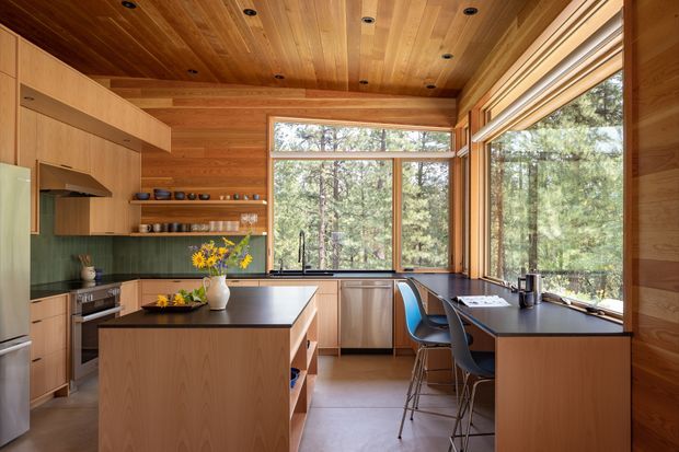 Warm, timber-lined contemporary kitchen with extensive corner glazing overlooking a wooded landscape. The room features a large central island with dark benchtop, integrated timber cabinetry, open shelving, a green vertical-tile splashback, stainless-steel appliances, and a built-in dining/work counter with stools. Recessed ceiling lights and natural timber surfaces create a cohesive, inviting interior.