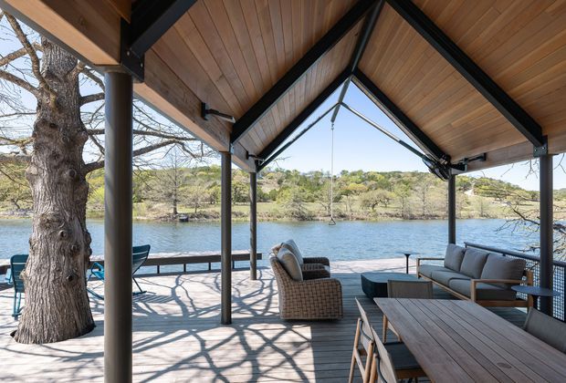 A covered waterfront outdoor living and dining terrace with a timber-lined pitched roof, exposed black structural beams, slender dark posts, and expansive open glazing. The space includes a large timber dining table, outdoor lounge seating, and woven armchairs overlooking a calm body of water and wooded shoreline. Bright natural daylight and warm timber surfaces create a relaxed, contemporary lakeside setting.