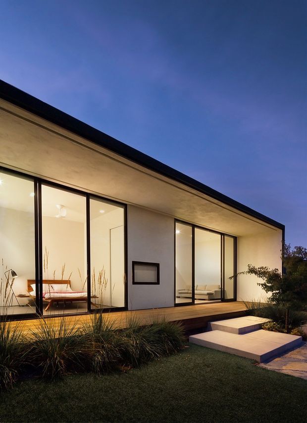 Modern single-storey residence viewed from a landscaped garden at dusk. The house has a long, low flat roof with dark trim, smooth light-coloured walls, expansive floor-to-ceiling glazed sliding doors with slim dark frames, and a timber deck. Warm interior lighting reveals a bedroom or living space through the glazing. Minimal concrete steps lead through a simple landscaped garden with grasses and shrubs.
