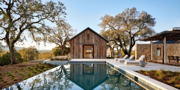 A landscaped outdoor pool courtyard overlooking a wooded, rolling landscape. The main feature is a long rectangular swimming pool reflecting a small rustic timber-clad gabled pavilion with an open doorway. To the right is a stone-clad single-storey structure with a covered outdoor dining or lounge area, while mature trees, planted garden beds, pale paving, and poolside loungers create a warm rural retreat atmosphere.