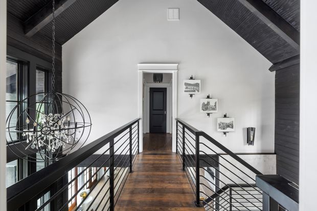 A high-level interior landing or hallway with a bridge-like walkway overlooking the lower level. The space features a steep vaulted white wall and dark exposed timber ceiling, dark timber flooring, black metal horizontal-bar balustrades, tall glazing on the left, and a sculptural spherical pendant light. A dark paneled door and small framed artworks sit at the far end, with stairs descending on the right.