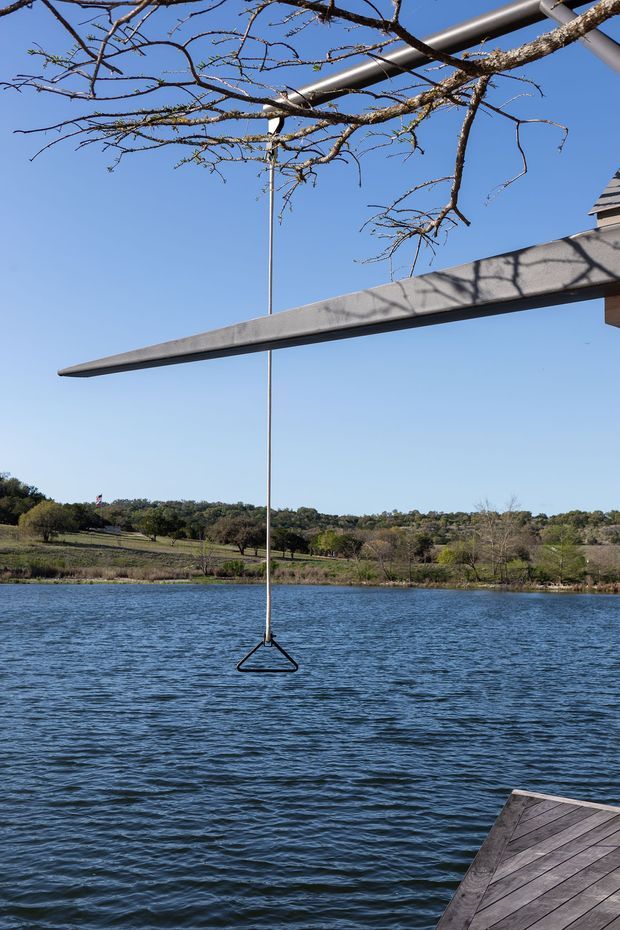 Outdoor lakeside view featuring a cantilevered grey concrete or metal beam structure extending over the water, with a suspended triangular swing or hanging element beneath it. A timber deck occupies the lower right corner, while leafless tree branches frame the upper portion and a rural, wooded shoreline lies across the lake.