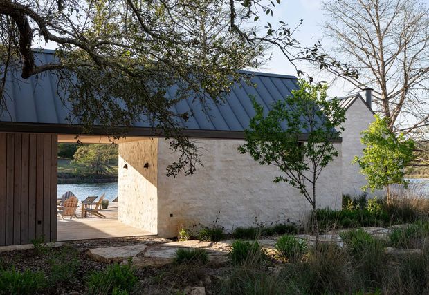 Exterior view of a low, contemporary structure with a dark standing-seam metal pitched roof, pale textured masonry or rendered walls, and a large timber-framed opening leading to a waterside terrace. Naturalistic grasses, shrubs, mature trees, and stone stepping elements surround the building. Outdoor seating and water are visible beyond the opening.