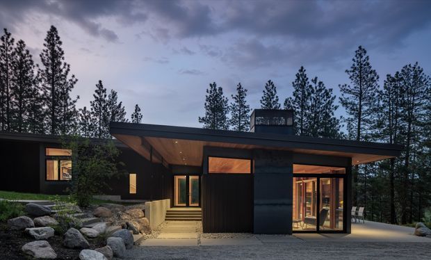 A low, single-storey modern house set among tall pine trees and rugged landscaped rocks at dusk. The dark-clad building has broad flat roof planes with deep overhangs, warm timber soffits, extensive floor-to-ceiling glazing, and a recessed entry reached by steps. Interior lighting creates a warm glow through the windows, contrasting with the cool blue evening sky.