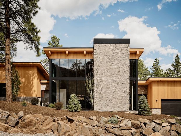 Contemporary single-storey residence set into a rocky, pine-covered landscape. The facade combines warm vertical timber cladding, expansive dark-framed floor-to-ceiling glazing, and a prominent pale stacked-stone central volume capped with a dark roof edge. A projecting timber-clad garage wing sits to the right, while exposed rock landscaping forms the foreground retaining edge.