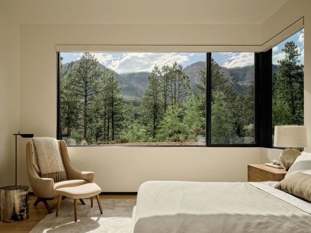 A calm, warmly lit bedroom with a large corner picture window framing a forested mountain landscape. The room features a neutral upholstered bed, mid-century-style lounge chair and ottoman, timber bedside table, table lamp, pale carpet, and dark window frames.