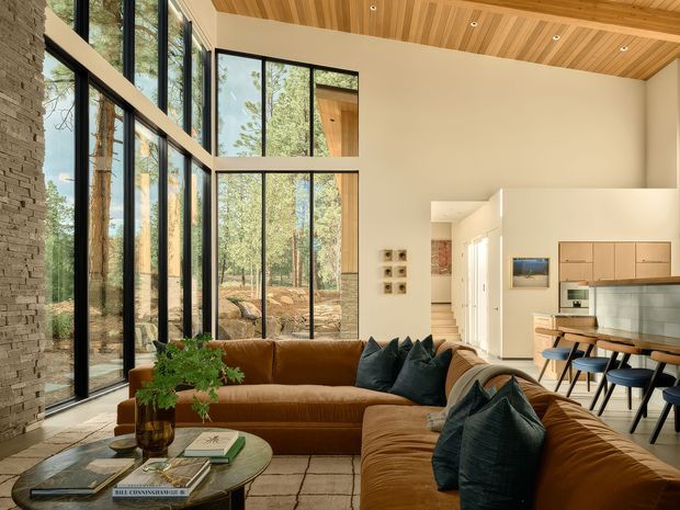 A refined open-plan living room with a large brown sectional sofa, dark cushions, a round coffee table, and a neutral rug. Floor-to-ceiling black-framed glazing brings a wooded landscape into view, while a warm timber slatted ceiling and stacked-stone feature wall add natural texture. The kitchen and dining area are visible beyond the living space, with pale cabinetry, a tiled backsplash, and a timber dining table.