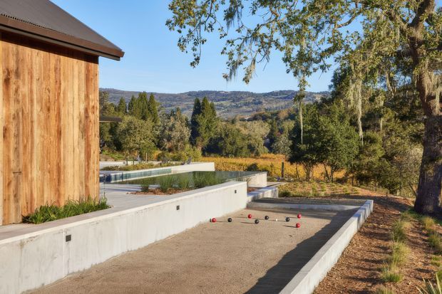 A landscaped outdoor recreation area beside a timber-clad building, featuring a long concrete-edged bocce court in the foreground and a partially visible swimming pool beyond. The setting has mature trees, dry natural planting, and expansive rolling hill views under clear daylight. The architecture combines warm vertical timber cladding with a dark pitched roof and restrained concrete landscape structures.