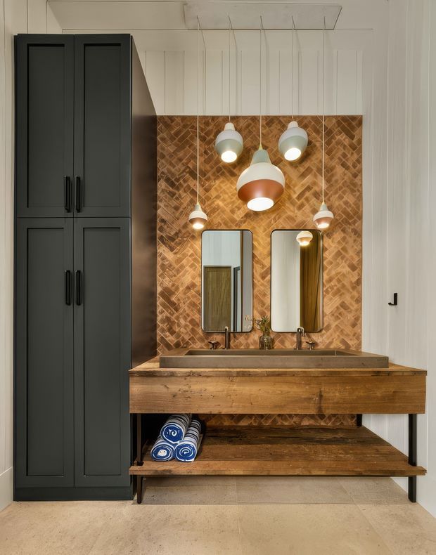 A stylish bathroom vanity wall with a large double timber vanity, two inset basins and brushed metal taps, backed by a warm herringbone timber feature wall. Two rounded mirrors sit above the basins, complemented by multiple suspended pendant lights. A tall matte-black paneled storage cabinet occupies the left side, while an open timber shelf below the vanity holds rolled towels. The room has light neutral flooring and white walls, creating a warm, refined contemporary look.