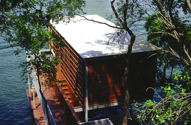 An elevated timber-clad structure projecting over a body of water, viewed through surrounding trees. The compact building has a dark timber screen wall, a bright low-pitched roof, and a narrow timber deck or walkway with simple metal supports and railings. The setting strongly suggests a waterfront or lakeside dwelling, cabin, or pavilion.