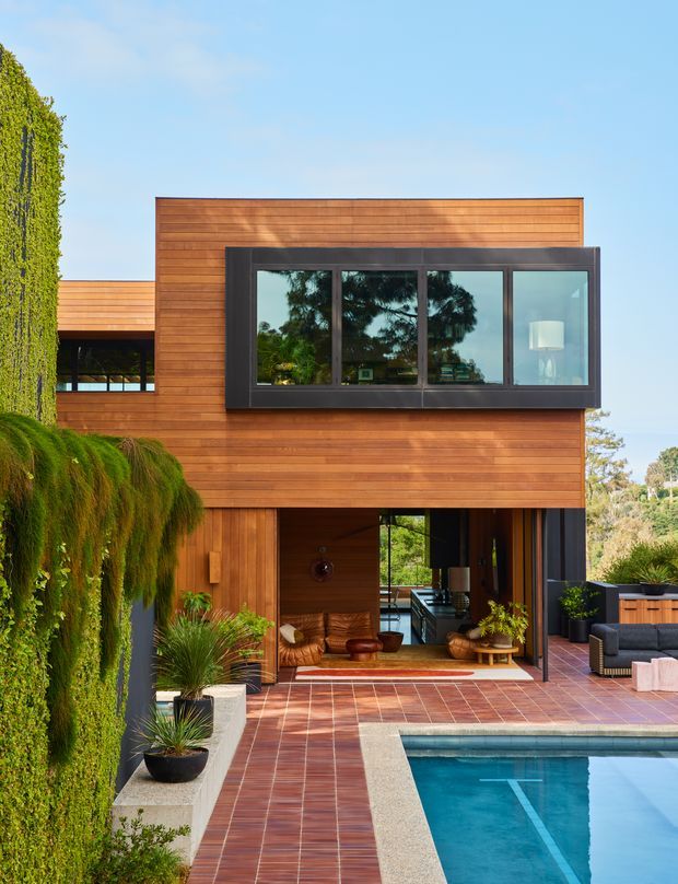 Exterior view of a contemporary two-storey home clad in horizontal warm timber boards, with a projecting dark-framed glazed window and an open ground-floor living area facing a tiled terrace and swimming pool. Lush vertical greenery and potted planting frame the scene, while outdoor lounge furniture is visible beside the pool.