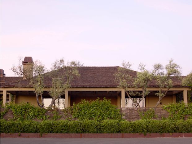 Single-storey residential building viewed from the street, with a long low tiled roof, chimney, warm earth-toned walls, deep covered veranda, timber or dark-framed openings, and landscaped hedges and mature trees screening the frontage. The architecture has a restrained traditional, Mediterranean-influenced appearance.