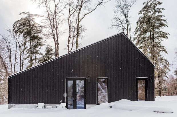 A simple black-clad gable-roofed building set in a snowy wooded landscape. The facade has vertical dark timber-like cladding, several narrow glazed doors or windows, and a minimal contemporary form.