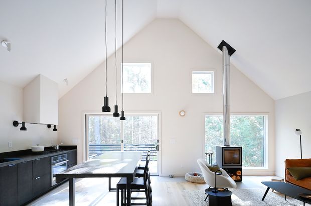 Bright open-plan living and dining space with a compact black kitchen at left, a long island with bar seating, large windows and glazed doors, and a freestanding wood-burning fireplace with a tall flue beneath a steeply pitched ceiling. Neutral finishes, pendant lights, contemporary furniture, and views of trees create an airy modern interior.