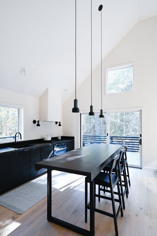 Bright contemporary kitchen and dining area with a large black island bench and integrated seating, matte black cabinetry, black pendant lights, pale timber flooring, and white walls. The room has a high sloping ceiling and generous glazing opening to a tree-filled exterior deck.