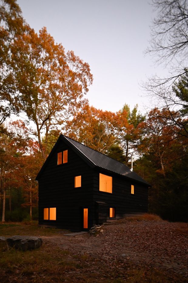 Exterior photograph of a compact, dark-clad cabin nestled among autumn trees. The two-storey building has a simple gable roof with dark corrugated metal roofing, black horizontal timber-like cladding, and several warm glowing rectangular windows and doors. A gravel and leaf-covered path leads toward the entrance, creating a secluded woodland setting at dusk.