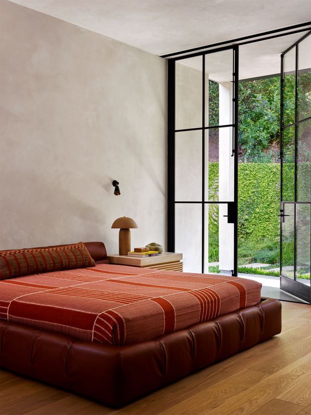 A warm, contemporary bedroom with a low upholstered leather bed in deep brown, layered with a rust-red striped bedspread and bolster cushion. The room has a softly textured plaster wall, pale concrete ceiling, timber flooring, a small timber bedside table with a sculptural lamp, and expansive black-framed steel-look glazed doors opening directly onto a manicured garden. The composition emphasizes the bed and the indoor-outdoor connection.
