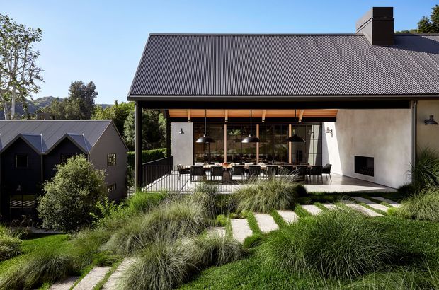 Exterior view of a contemporary dark-roofed house on a landscaped sloping site. The building features a large covered outdoor entertaining terrace with black-framed glazing, exposed timber elements, dining furniture, and an outdoor fireplace integrated into a pale rendered wall. A smaller dark-clad gabled building is visible to the side, surrounded by ornamental grasses and stepping-stone paths.