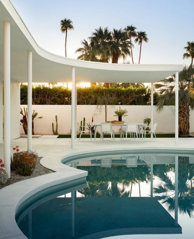 A bright outdoor pool courtyard with a curved white shade canopy supported by slender columns. The pool has a rounded, organic edge and reflects the canopy and surrounding palm trees. White outdoor dining furniture sits on a pale paved terrace, bordered by white rendered walls, clipped hedges, desert plants, cacti and mature palms. The setting has a clean, resort-like modernist character with strong mid-century influences.