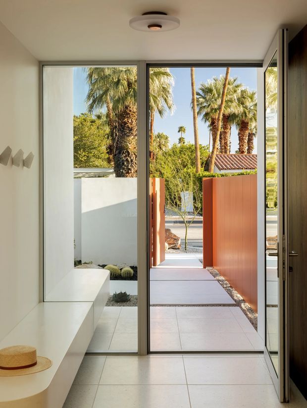 A bright minimalist interior entryway opens directly onto a landscaped courtyard and walkway. The space features smooth white built-in bench seating, pale tiled flooring, white walls, full-height dark-framed glazing and a timber door. Outside, a terracotta privacy screen, gravel garden beds, palms and a low tiled roof create a warm desert-modern setting.
