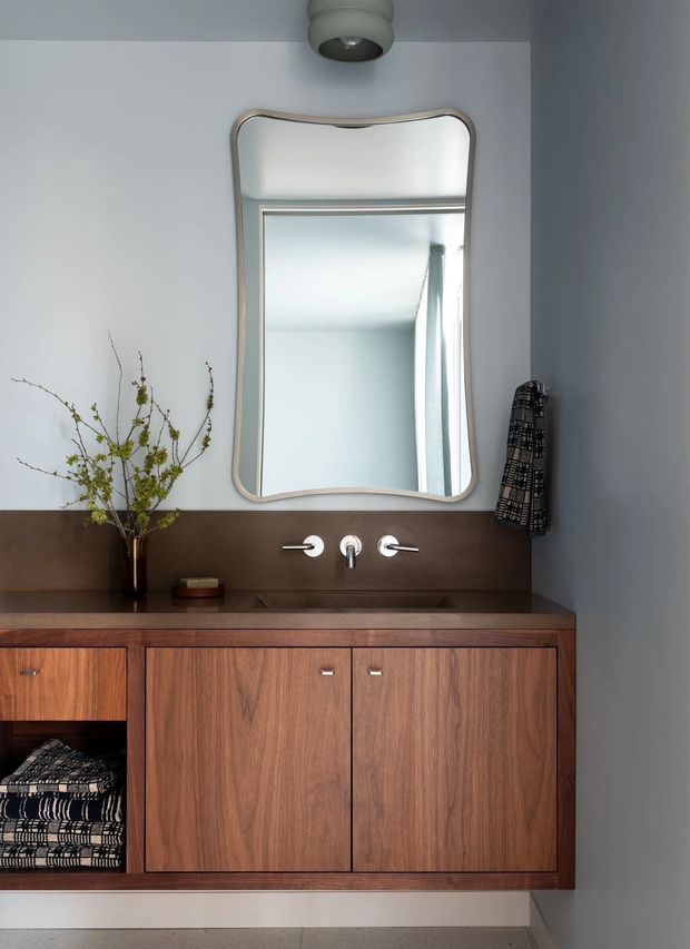 A refined bathroom vanity area with a floating dark timber cabinet, integrated open shelving, dark brown benchtop and backsplash, wall-mounted chrome mixer tap, and a large organically shaped framed mirror. A sculptural ceiling light and minimal styling complete the warm, contemporary composition.