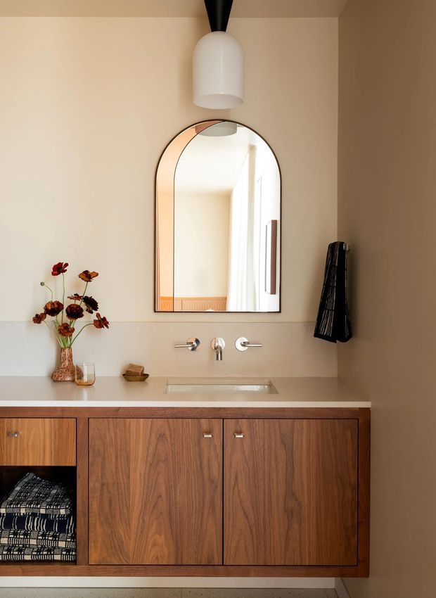 A warm, refined bathroom vanity with a floating or recessed walnut-toned timber cabinet, pale stone benchtop and integrated basin. Brushed metal wall-mounted tapware sits beneath a tall arched mirror with a dark frame. A sculptural white pendant light, dried floral arrangement and black textile add decorative accents. The room has soft beige walls and a calm, contemporary character.