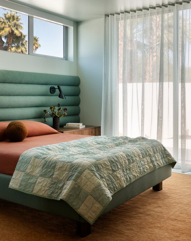 A softly lit bedroom with a low upholstered bed, a distinctive rounded horizontal-panel green headboard, terracotta bedding, and a pale green-blue quilt. Floor-to-ceiling sheer curtains cover large windows and doors, with palm trees visible outside. A timber bedside cabinet, small wall-mounted reading light, and minimal bedside decor complete the warm, calm interior.