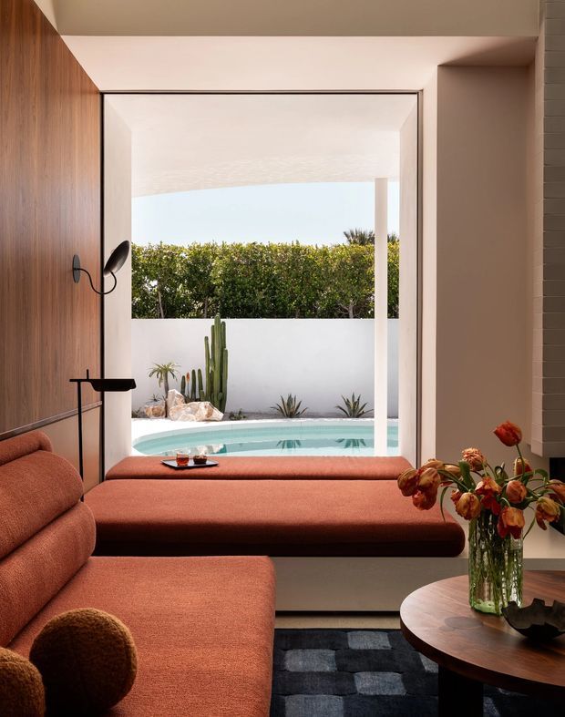 A warm, sophisticated living room framed around a large open glazed opening to a private courtyard pool. The room features a substantial rust-orange sectional sofa and matching built-in daybed, a dark timber wall panel, slim black floor and wall lighting, a round timber coffee table, patterned dark rug, and floral arrangement. Outside are a turquoise swimming pool, white rendered walls, sculptural cacti, and dense screening greenery. The composition has strong indoor-outdoor flow and a refined contemporary feel.