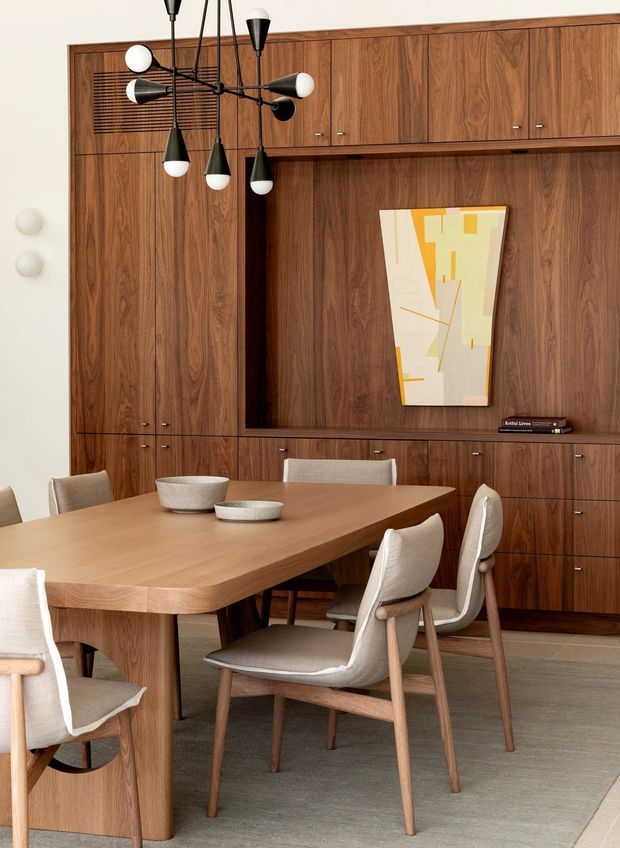 Warmly styled dining room with a large mid-century-inspired timber dining table and upholstered wooden dining chairs. The room is dominated by a full-height dark timber built-in wall with extensive cabinetry, a recessed display niche, artwork, and integrated louvred detailing. A sculptural black-and-white multi-arm pendant hangs above the table, while neutral walls, flooring, and upholstery create a refined, earthy palette.
