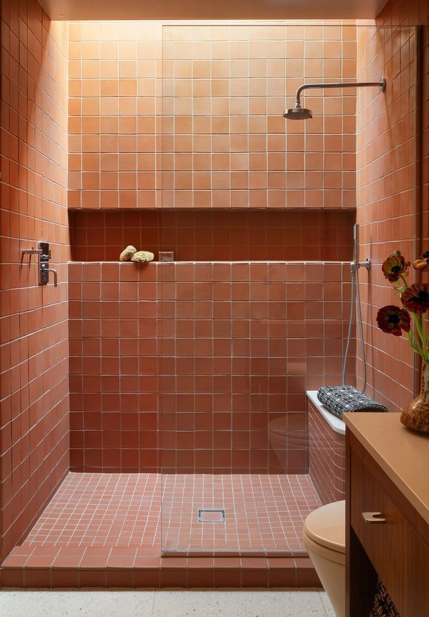 A compact walk-in shower bathroom clad almost entirely in small terracotta-pink square tiles, including the walls, built-in recessed shelf, shower floor, and raised threshold. The shower has a ceiling-height tiled enclosure, a dark metal rainfall showerhead and handheld fitting, a built-in tiled bench with a patterned cushion, and a partial view of a toilet and timber vanity at right. Warm lighting and terracotta surfaces create a distinctive retro, intimate atmosphere.