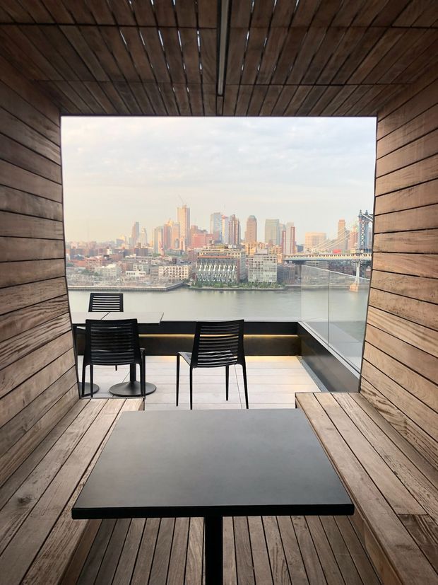A compact, wood-lined balcony or terrace framed like an outdoor room, with vertical timber wall and ceiling boards, a dark rectangular dining table, and black outdoor chairs. Full-height glass balustrades open to a prominent riverside city skyline and bridge beyond. The space has a warm, contemporary architectural feel and appears elevated above the water.