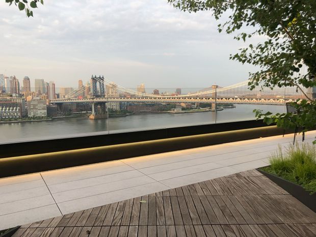 A landscaped waterfront terrace overlooking a wide river, suspension bridges, and a dense urban skyline. The foreground features a warm timber deck, large pale paving slabs, integrated linear lighting, dark metal privacy or safety screening, and raised planters with ornamental grasses and trees. The scene has a refined contemporary public-terrace character.