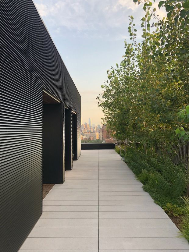 A contemporary rooftop terrace walkway beside a dark, louvred-clad building. The terrace has large pale rectangular paving slabs, black metal railings, dense planted greenery, and a distant urban skyline visible at the end of the path. The scene has clean geometric lines and a calm, modern outdoor atmosphere.