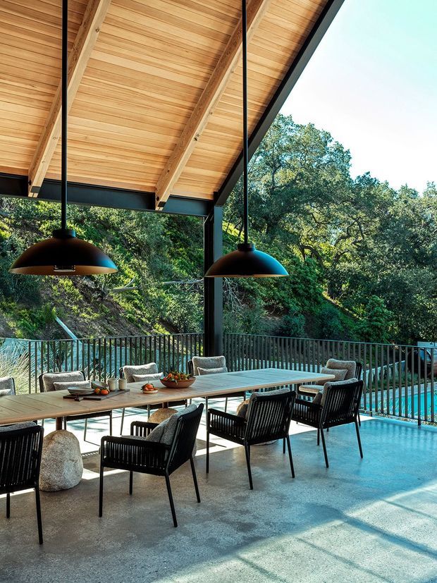 A covered outdoor dining terrace with a long timber dining table and black woven armchairs, overlooking a turquoise swimming pool and wooded landscape. The space has a broad timber-lined pitched roof with exposed rafters, dark structural steel beams and columns, pendant lighting, and a concrete floor, creating a refined indoor-outdoor entertaining area.