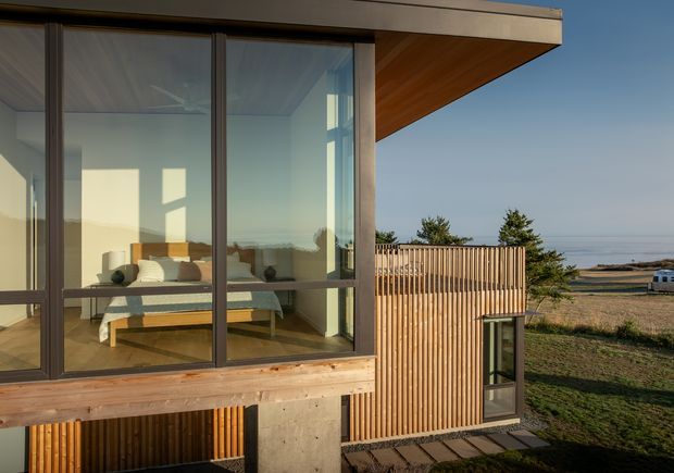 Exterior view of a contemporary coastal residence with extensive floor-to-ceiling glazing, dark-framed sliding doors, warm timber soffit and horizontal timber base cladding, vertical timber screening, and exposed concrete elements. A bedroom is clearly visible through the glazing, while the building overlooks a dry coastal landscape and ocean horizon.