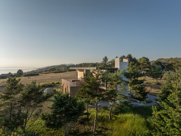 A modern multi-level house set within a natural coastal landscape, viewed from an elevated distance. The building has a compact rectilinear form, extensive glazing, timber-clad sections, and a prominent pale masonry or concrete vertical tower. Native-looking trees and grasses surround the home, with an open shoreline or water visible in the distance.