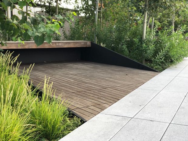A landscaped outdoor terrace featuring a broad dark timber deck set slightly below a pale large-format paved walkway. A black rendered retaining wall and low timber-edged planter border the deck, with dense green planting and ornamental grasses surrounding the space. The scene has a clean, contemporary garden design with strong geometric lines and layered greenery.