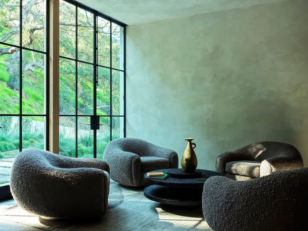 A moody, contemporary living room with sculptural boucle lounge chairs and sofas arranged around layered black oval coffee tables. A large corner of black-framed floor-to-ceiling glazing brings in abundant natural light and views of lush greenery, while textured pale green-grey plaster walls and a concrete-like ceiling create a calm, organic atmosphere.