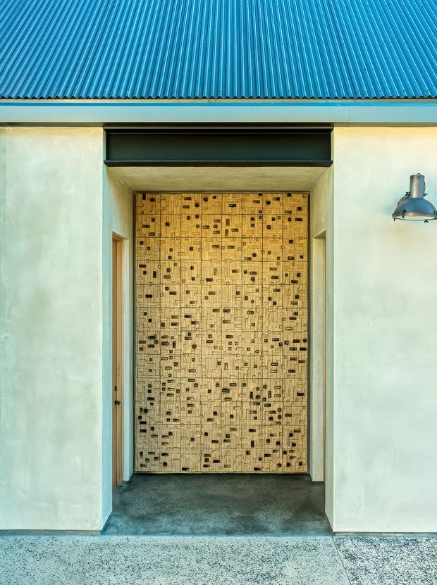 Front-facing close view of a minimalist building entry. A large patterned timber door with numerous small dark inlays sits within a recessed pale plaster surround, beneath a dark horizontal transom or louvre opening. The facade has a vivid blue corrugated roof, a simple exterior wall-mounted light, and a dark polished threshold with speckled paving in front.