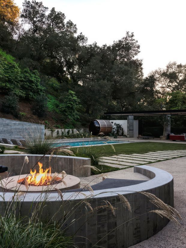 A landscaped outdoor entertaining area set against a wooded hillside, featuring a swimming pool, a curved concrete fire-pit seating structure with an active fire, ornamental grasses, stepping-stone paving, and a covered pavilion with a dark flat roof and concrete supports. A cylindrical outdoor feature is visible beside the pavilion.