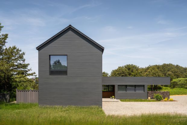 Exterior view of a contemporary single-storey residential building with a tall gabled volume clad in dark horizontal weatherboards and a lower flat-roofed wing. Dark-framed windows, a pale gravel forecourt, timber fencing, landscaped planting, and mature trees surround the house.