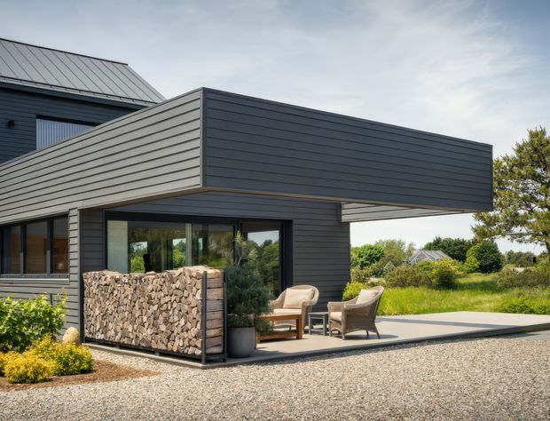Exterior view of a contemporary dark-clad house with a projecting box-like upper volume and covered patio. Horizontal siding, a standing-seam metal gable roof, large glazed openings, and a built-in firewood storage wall define the facade. Outdoor lounge chairs sit on a paved terrace overlooking a landscaped garden and gravel foreground.